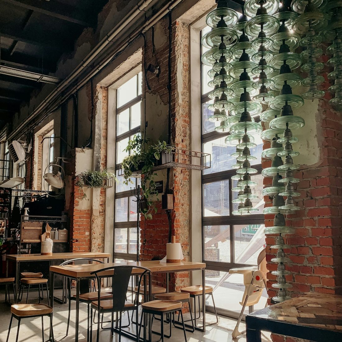 Bright, plant-filled café interior with exposed brick walls and hanging glass pendant lights over wooden tables