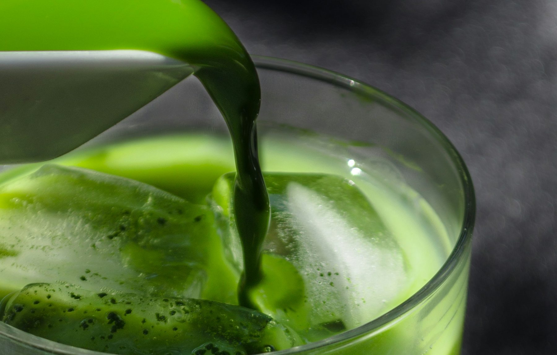 Bright green matcha being poured over ice into a glass, steam rising against a dark background